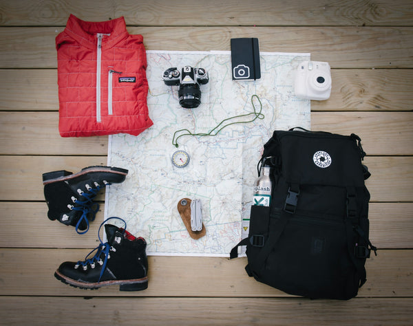 A flat-lay of outdoor gear including a hiking backpack, leather boots, a jacket, map, and compass laid out for cleaning and packing