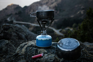 A blue and gray camping gas burner with a cooking pan set up on a cliff at dusk