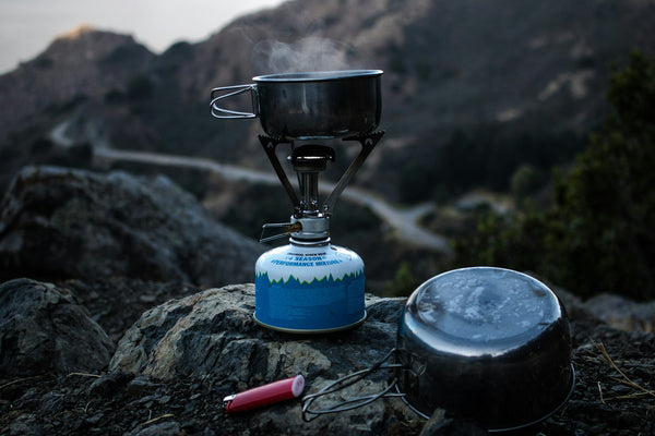 A blue and gray camping gas burner with a cooking pan set up on a cliff at dusk