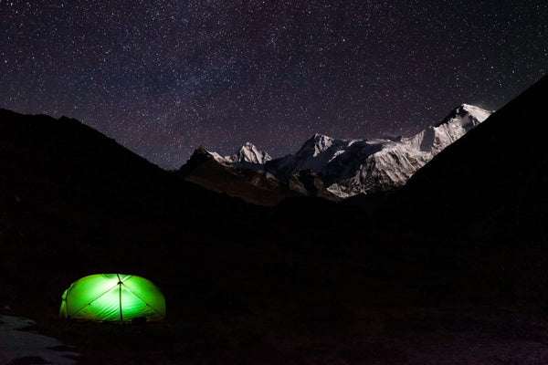 A glowing tent at a campsite beneath snowy mountain peaks and a starry night sky